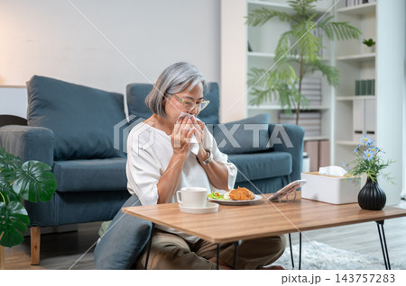 Senior Asian woman wiping mouth with napkin while eating at low table in a living room. 143757283
