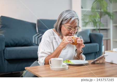 Senior Asian woman biting into croissant held with both hands at table in a living room. Senior Asian woman biting into croissant held with both hands at table in a living room. 143757285