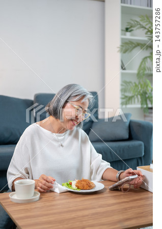 Senior Asian woman smiling while looking at phone near breakfast on table in a living room. 143757286