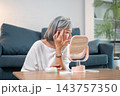 Elderly asian woman inspecting crow feet and eye wrinkles in a mirror on table. 143757350