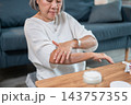 Senior asian woman gently spreading body moisturizer on arm while sitting on living room floor. 143757355