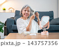 Senior asian woman combing or brushing her bob grey hair while looking in mirror at a living room 143757356
