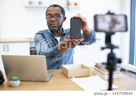 A man presents a smartphone at a desk while recording a video, with a laptop, open parcel box, stacked books, and camera equipment in a bright workspace A man presents a smartphone at a desk while recording a video, with a laptop, open parcel box, stacked books, and camera equipment in a bright workspace 143759044