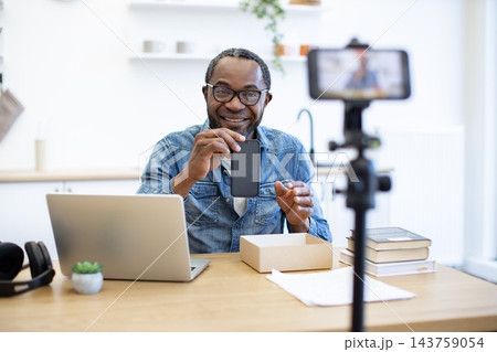 A smiling creator records a desk-side video with a smartphone, laptop, books, and product box in a bright, organized home workspace 143759054