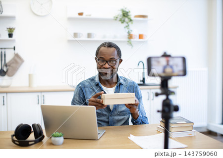An African American man presents a small cardboard package to a smartphone camera from a bright home workspace, with a laptop and books nearby 143759060