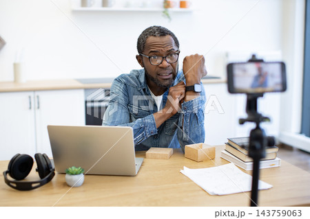 A man presents a smartwatch to a phone camera at a home desk, with a laptop, headphones, books, and shipping boxes arranged around his workspace 143759063