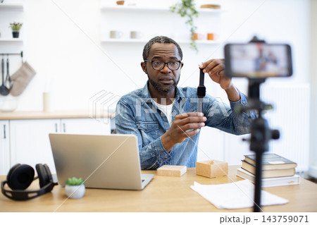 A man presents a smartwatch at a desk while recording with a smartphone, with a laptop, headphones, books, papers, and small wooden blocks nearby A man presents a smartwatch at a desk while recording with a smartphone, with a laptop, headphones, books, papers, and small wooden blocks nearby 143759071