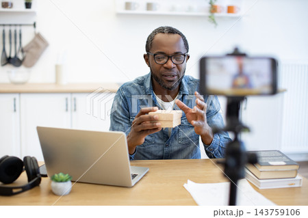 He addresses a mounted phone while holding a small box, using a laptop at a desk in a bright home kitchen with books and headphones nearby He addresses a mounted phone while holding a small box, using a laptop at a desk in a bright home kitchen with books and headphones nearby 143759106