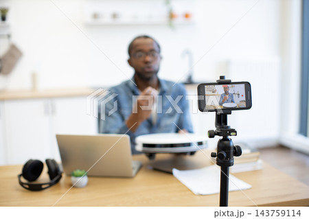A creator records a hands-on robot vacuum review in a bright kitchen, with a smartphone on a tripod capturing the demonstration beside a laptop A creator records a hands-on robot vacuum review in a bright kitchen, with a smartphone on a tripod capturing the demonstration beside a laptop 143759114