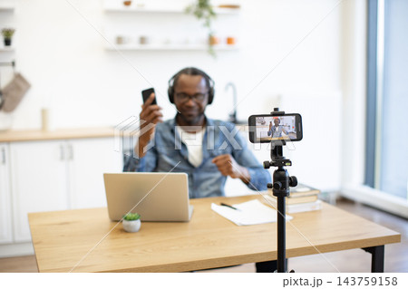 A man wearing headphones speaks toward a smartphone mounted on a tripod while seated at a laptop in a bright home kitchen workspace with papers and a small potted plant nearby A man wearing headphones speaks toward a smartphone mounted on a tripod while seated at a laptop in a bright home kitchen workspace with papers and a small potted plant nearby 143759158