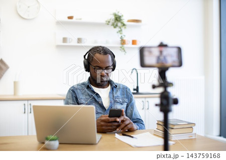 A man works at a kitchen table with a laptop and phone as a smartphone on a tripod captures him in a bright, uncluttered home setting A man works at a kitchen table with a laptop and phone as a smartphone on a tripod captures him in a bright, uncluttered home setting 143759168