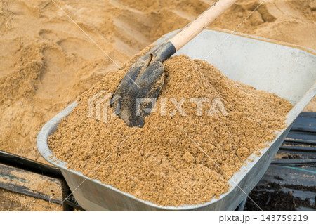 A sturdy shovel leans against a metal wheelbarrow that is filled with fine sand. The scene captures essential tools used for manual labor at a busy construction site. 143759219