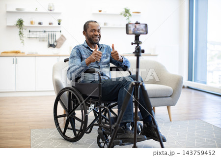 A smiling man gives two thumbs up while seated in a wheelchair, recording a video on a smartphone in a bright, modern living room at home 143759452