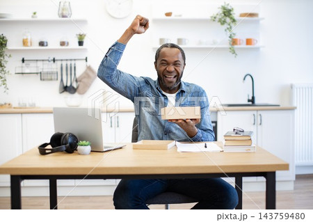 A joyful man raises his fist while holding a small box at a kitchen table, with a laptop, books, headphones, and papers in a bright home setting A joyful man raises his fist while holding a small box at a kitchen table, with a laptop, books, headphones, and papers in a bright home setting 143759480