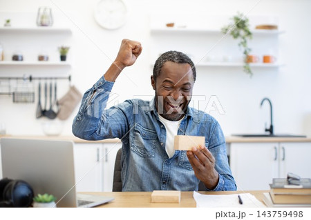 A joyful African American man raises a fist while holding a small parcel at his desk, with a laptop, paperwork, and a bright kitchen behind him 143759498