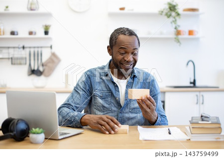 An African American man smiles while examining a small cardboard package at his desk, with a laptop, headphones, notebook, and books in a bright workspace 143759499