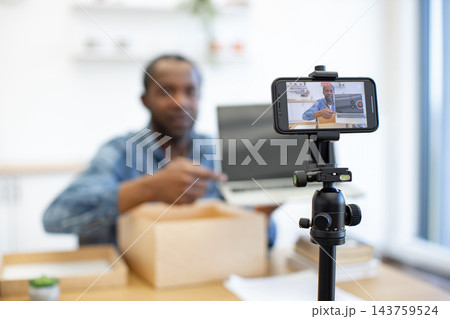 A man films himself with a smartphone on a tripod while opening a cardboard package beside a laptop in a bright home workspace at a desk A man films himself with a smartphone on a tripod while opening a cardboard package beside a laptop in a bright home workspace at a desk 143759524