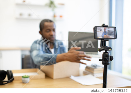 A creator demonstrates a cardboard package at a desk, filming with a smartphone on a tripod beside a laptop in a bright home workspace A creator demonstrates a cardboard package at a desk, filming with a smartphone on a tripod beside a laptop in a bright home workspace 143759525