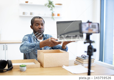 An African American man presents a laptop to a smartphone camera, with cardboard packaging, books, and desk accessories in a bright workspace An African American man presents a laptop to a smartphone camera, with cardboard packaging, books, and desk accessories in a bright workspace 143759526