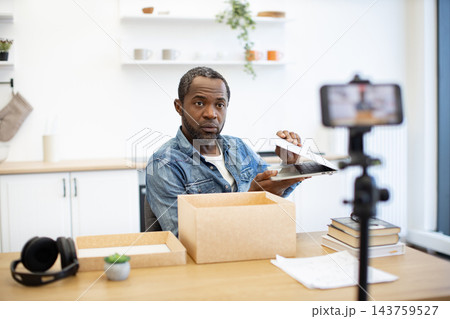 A content creator records a laptop unboxing at a bright home workspace, with cardboard packaging, books, headphones, and a phone mounted on a tripod A content creator records a laptop unboxing at a bright home workspace, with cardboard packaging, books, headphones, and a phone mounted on a tripod 143759527