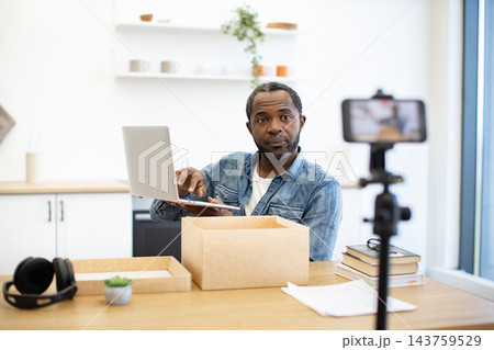 An African American man works at a desk with a laptop and shipping boxes while a smartphone on a tripod records his home workspace for online content 143759529
