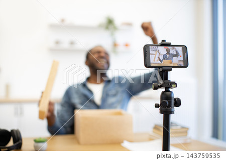 A smiling Black creator films a hands-on demonstration at a worktable, with a smartphone on a tripod, cardboard box, and wooden piece in view A smiling Black creator films a hands-on demonstration at a worktable, with a smartphone on a tripod, cardboard box, and wooden piece in view 143759535