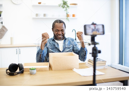 An African American man celebrates at a home office desk with raised fists beside a cardboard box, headphones, books, and a phone on a tripod An African American man celebrates at a home office desk with raised fists beside a cardboard box, headphones, books, and a phone on a tripod 143759540