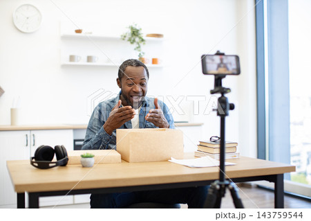 An African American man gestures toward an open cardboard package while recording a video at a bright home desk with a smartphone on a tripod 143759544