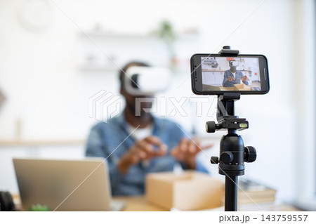 A creator wearing a virtual reality headset demonstrates a hands-on activity while a smartphone records the scene in a bright home workspace A creator wearing a virtual reality headset demonstrates a hands-on activity while a smartphone records the scene in a bright home workspace 143759557