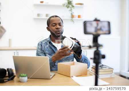 An African American creator examines a virtual reality headset at a desk, with a laptop, recording phone, shipping box, books and studio equipment nearby An African American creator examines a virtual reality headset at a desk, with a laptop, recording phone, shipping box, books and studio equipment nearby 143759570