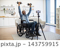 A smiling man in a wheelchair lifts dumbbells at home while recording his adaptive workout with a smartphone mounted on a tripod in a bright living space 143759915