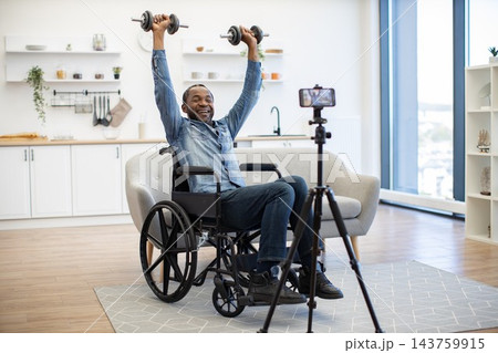 A smiling man in a wheelchair lifts dumbbells at home while recording his adaptive workout with a smartphone mounted on a tripod in a bright living space 143759915
