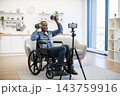 A smiling man in a wheelchair lifts dumbbells and records his workout with a smartphone on a tripod in a bright, contemporary living room 143759916