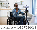 A smiling man in a wheelchair lifts dumbbells while recording a home workout, highlighting accessible fitness and independent strength training 143759918