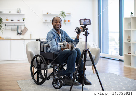 A smiling man builds upper-body strength from his wheelchair while recording a home workout with a smartphone mounted on a tripod in a bright living space A smiling man builds upper-body strength from his wheelchair while recording a home workout with a smartphone mounted on a tripod in a bright living space 143759921