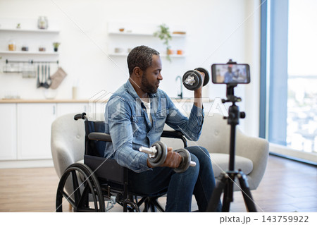 A man in a wheelchair lifts dumbbells at home while a smartphone on a tripod records his strength training session beside a bright window A man in a wheelchair lifts dumbbells at home while a smartphone on a tripod records his strength training session beside a bright window 143759922