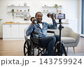 A smiling African American man lifts dumbbells from his wheelchair while a smartphone on a tripod records his strength workout in a bright living room 143759924