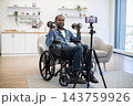 A smiling man exercises from his wheelchair in a bright modern home, lifting weights while recording a fitness session with a phone on a tripod 143759926