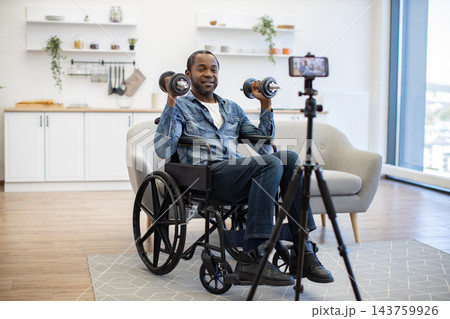 A smiling man exercises from his wheelchair in a bright modern home, lifting weights while recording a fitness session with a phone on a tripod 143759926