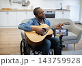 An African American musician plays acoustic guitar from his wheelchair in a bright home, while a smartphone on a tripod records the performance 143759928