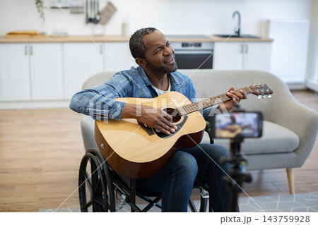 An African American musician plays acoustic guitar from his wheelchair in a bright home, while a smartphone on a tripod records the performance 143759928