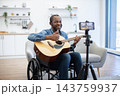 A smiling man plays acoustic guitar while recording a video at home, seated in a wheelchair in a bright living room with a phone on tripod 143759937