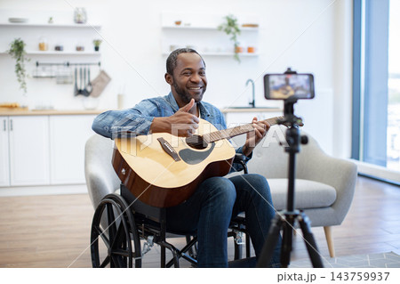A smiling man plays acoustic guitar while recording a video at home, seated in a wheelchair in a bright living room with a phone on tripod A smiling man plays acoustic guitar while recording a video at home, seated in a wheelchair in a bright living room with a phone on tripod 143759937