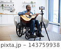 A smiling musician plays acoustic guitar from his wheelchair while recording a home performance on a smartphone in a bright modern living room 143759938