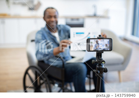 A man in a wheelchair demonstrates a boxed item while filming himself with a smartphone mounted on a tripod in a bright, modern indoor setting 143759940