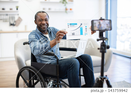 A smiling professional presents a rising chart to a phone camera from his wheelchair in a bright home office, sharing business results with viewers online 143759942