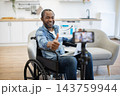 A smiling wheelchair user presents a rising chart and gives a thumbs-up while working at a laptop, with a phone mounted on a tripod nearby 143759944