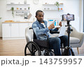 A wheelchair user presents a printed chart to a camera while working on a laptop in a bright home kitchen with a recording setup nearby 143759957