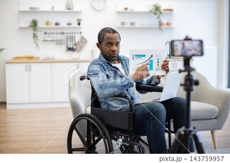 A wheelchair user presents a printed chart to a camera while working on a laptop in a bright home kitchen with a recording setup nearby 143759957