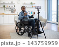 An African American presenter shares a printed business chart from his wheelchair while a smartphone on a tripod records him in a home interior 143759959